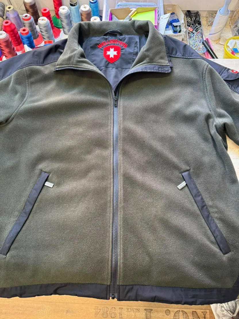 Eine graue Fleecejacke mit durchgehendem Reißverschluss und einem kleinen Emblem am Kragen, flach auf dem Arbeitstisch.