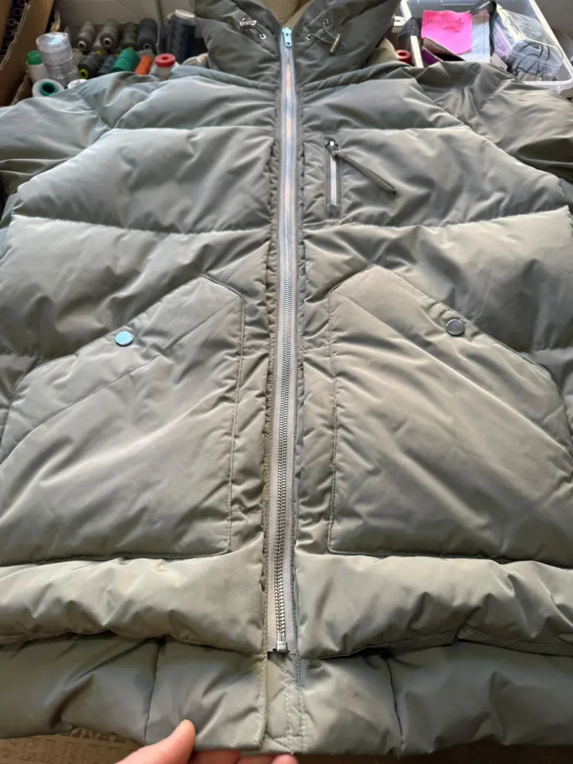 Eine graue Steppjacke liegt flach mit geöffnetem Reißverschluss, im Hintergrund die Wand mit den Garnrollen der Werkstatt.