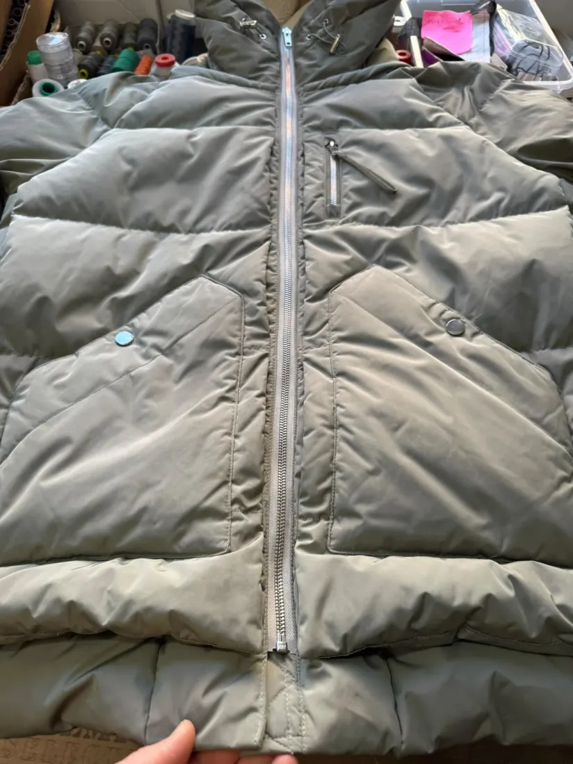 Eine graue Steppjacke liegt flach mit geöffnetem Reißverschluss, im Hintergrund die Wand mit den Garnrollen der Werkstatt.