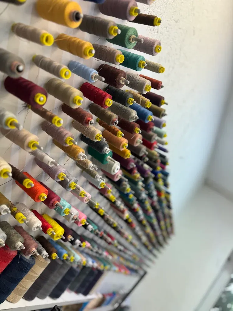 Eine Wand mit mehreren hundert Garnrollen, waagerecht auf Stifte gesteckt und nach Farbtönen gruppiert: oben Gelb und Ocker, darunter Rot, Grün, Blau, Rosa und Grau. Die Aufnahme läuft schräg an der Wand entlang, sodass die Reihen sich nach hinten verlieren.